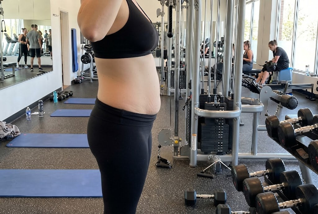 Mujer de perfil en un gimnasio, con el vientre posparto visible sobre las mallas